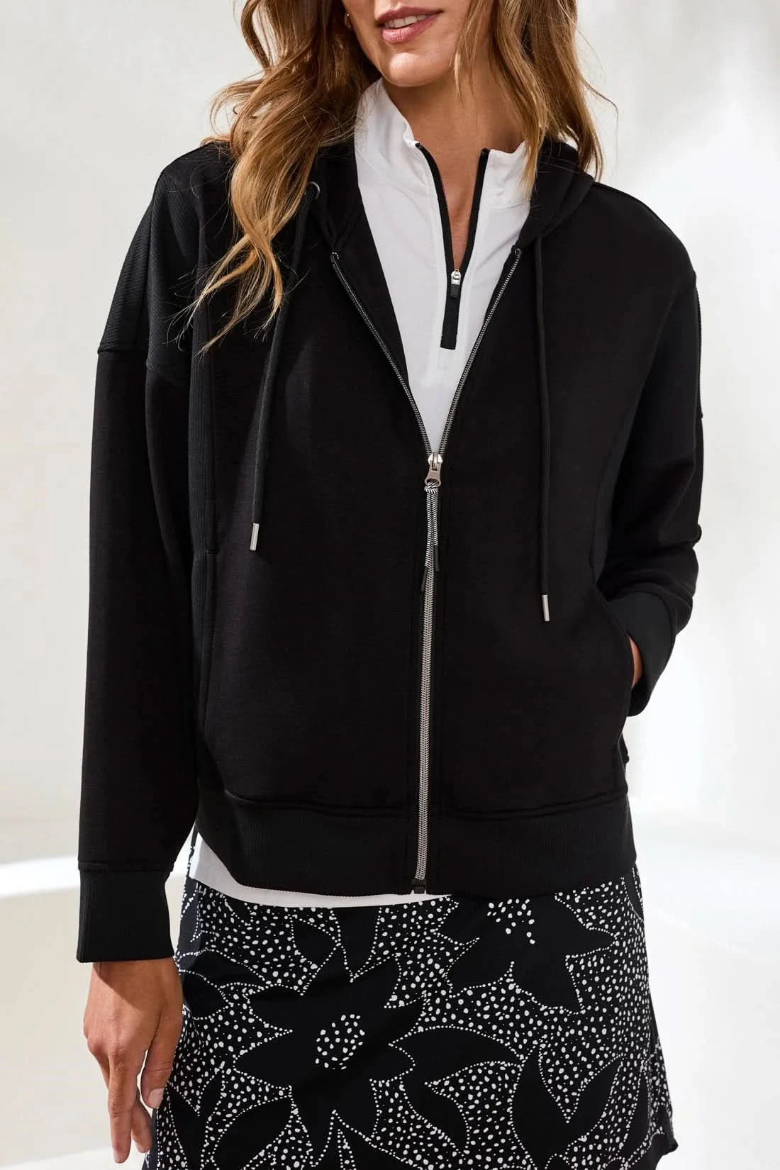 A woman stands indoors wearing the Tribal TECHNO LUX HOODED CARDIGAN COMBO, a black stretch scuba knit full-zip sweatshirt, over a white collared shirt and patterned black-and-white pants, with her hands in the cardigan pockets.