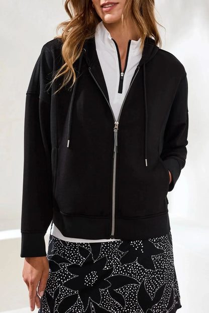 A woman stands indoors wearing the Tribal TECHNO LUX HOODED CARDIGAN COMBO, a black stretch scuba knit full-zip sweatshirt, over a white collared shirt and patterned black-and-white pants, with her hands in the cardigan pockets.