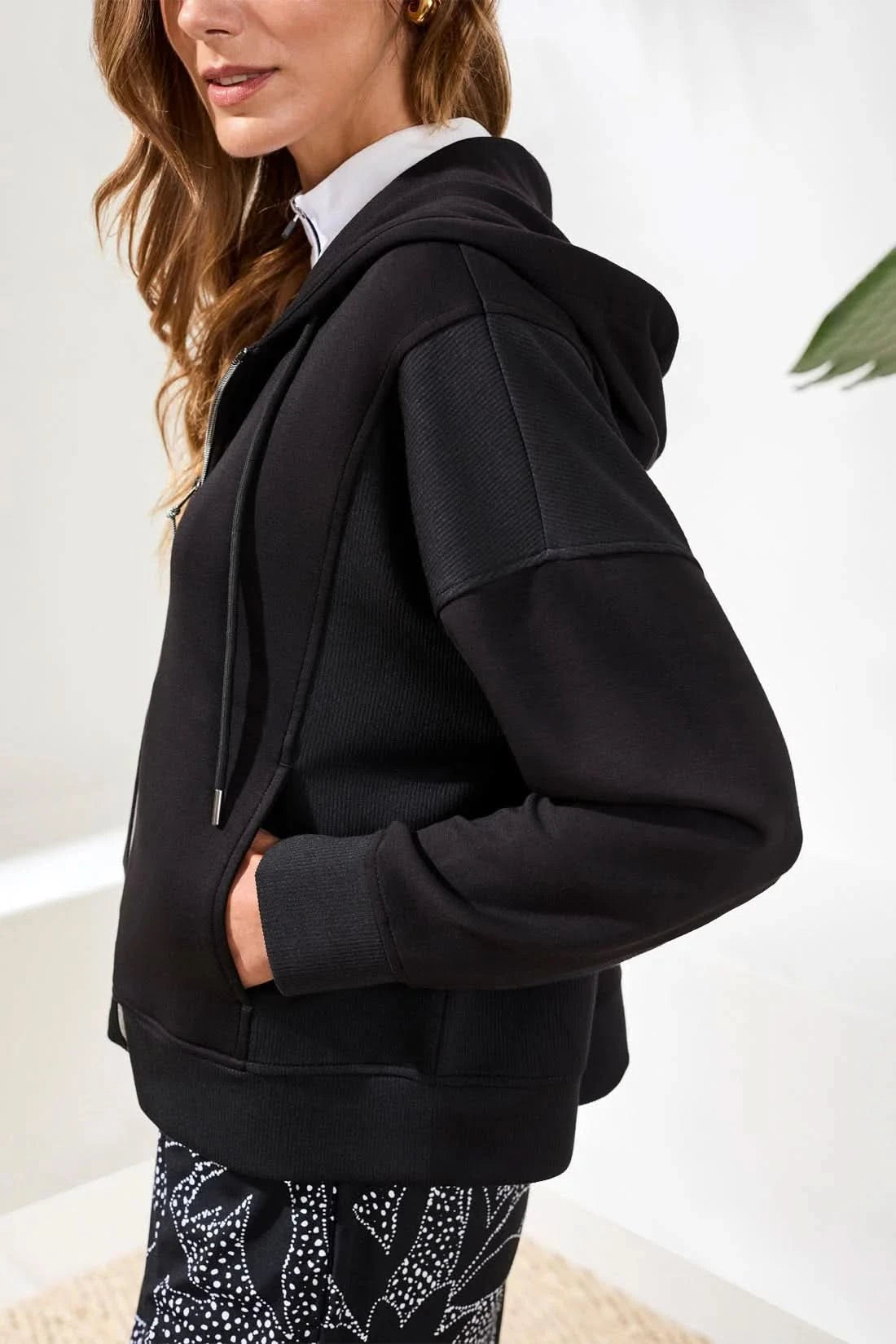 A woman stands indoors wearing the Tribal TECHNO LUX HOODED CARDIGAN COMBO, a black stretch scuba knit full-zip sweatshirt, over a white collared shirt and patterned black-and-white pants, with her hands in the cardigan pockets.