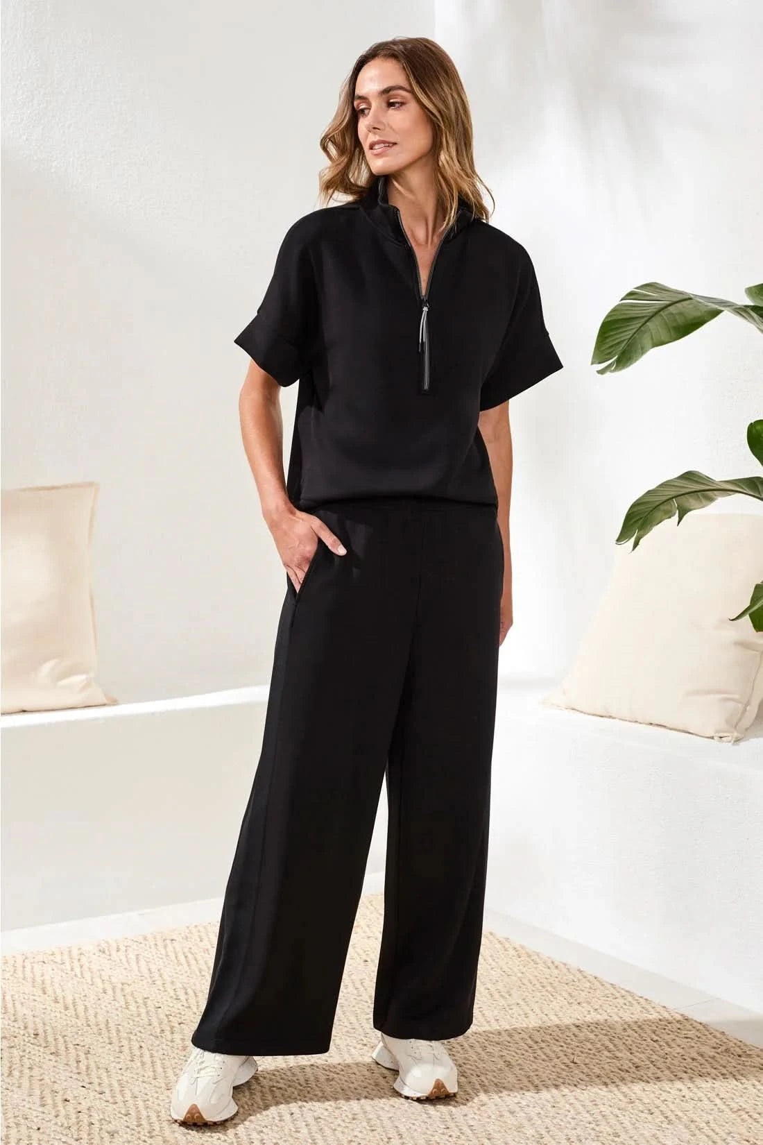 A woman in Tribal’s TECHNO LUX LOUNGE PANT, a black top, and white sneakers stands indoors by a white sofa with a beige cushion and green plant—showcasing effortless women’s activewear style.