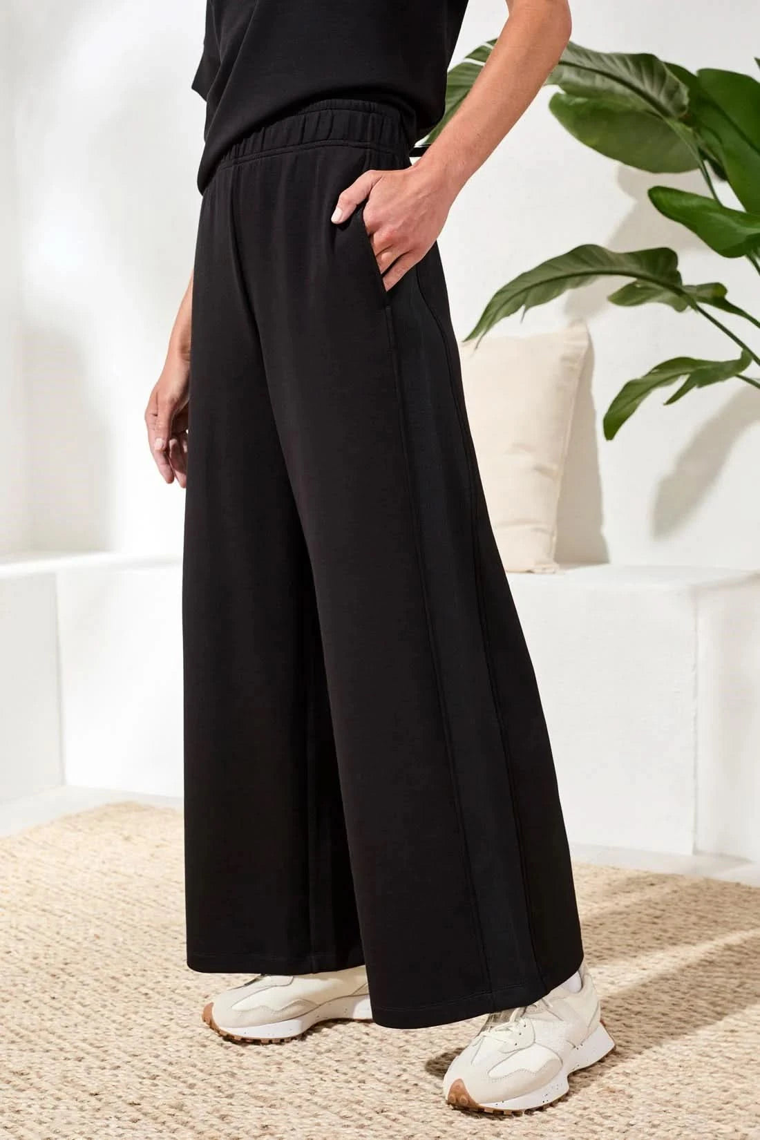 A woman in Tribal’s TECHNO LUX LOUNGE PANT, a black top, and white sneakers stands indoors by a white sofa with a beige cushion and green plant—showcasing effortless women’s activewear style.