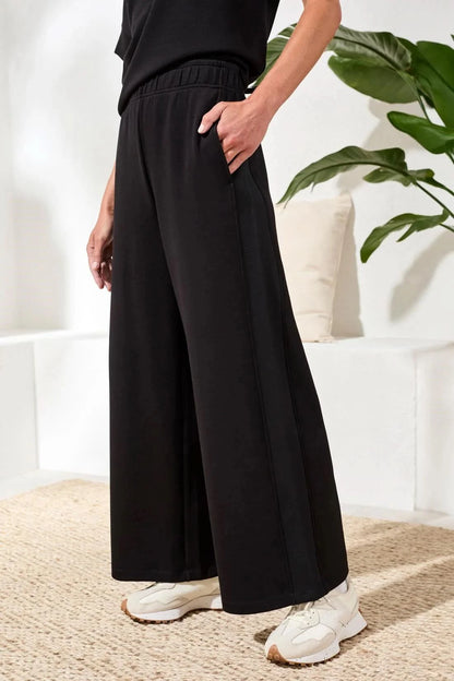 A woman in Tribal’s TECHNO LUX LOUNGE PANT, a black top, and white sneakers stands indoors by a white sofa with a beige cushion and green plant—showcasing effortless women’s activewear style.
