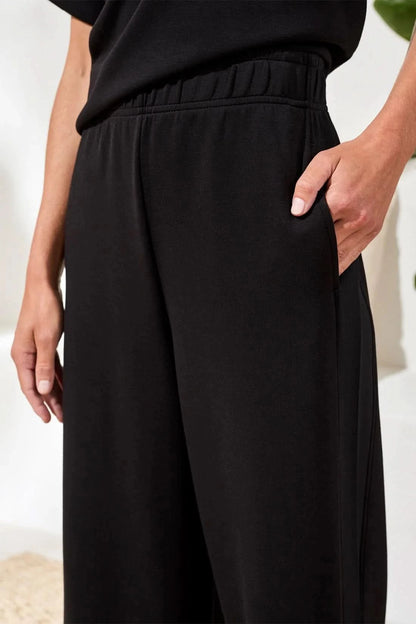 A woman in Tribal’s TECHNO LUX LOUNGE PANT, a black top, and white sneakers stands indoors by a white sofa with a beige cushion and green plant—showcasing effortless women’s activewear style.