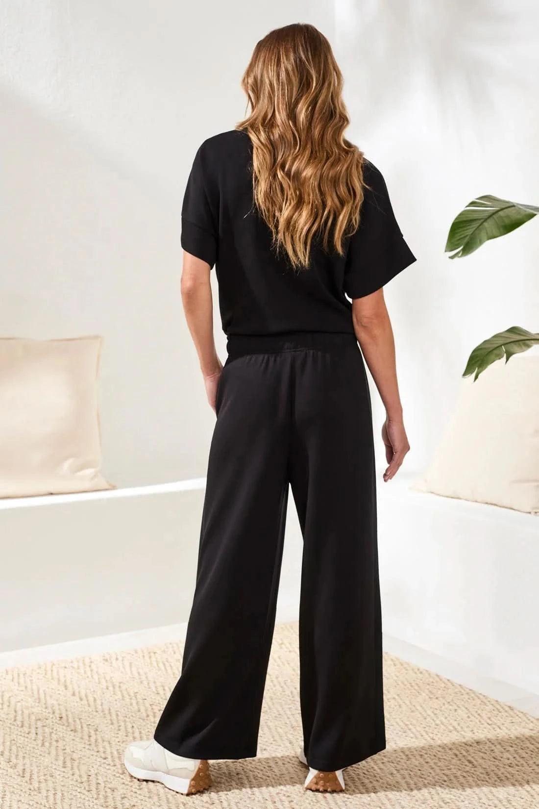 A woman in Tribal’s TECHNO LUX LOUNGE PANT, a black top, and white sneakers stands indoors by a white sofa with a beige cushion and green plant—showcasing effortless women’s activewear style.