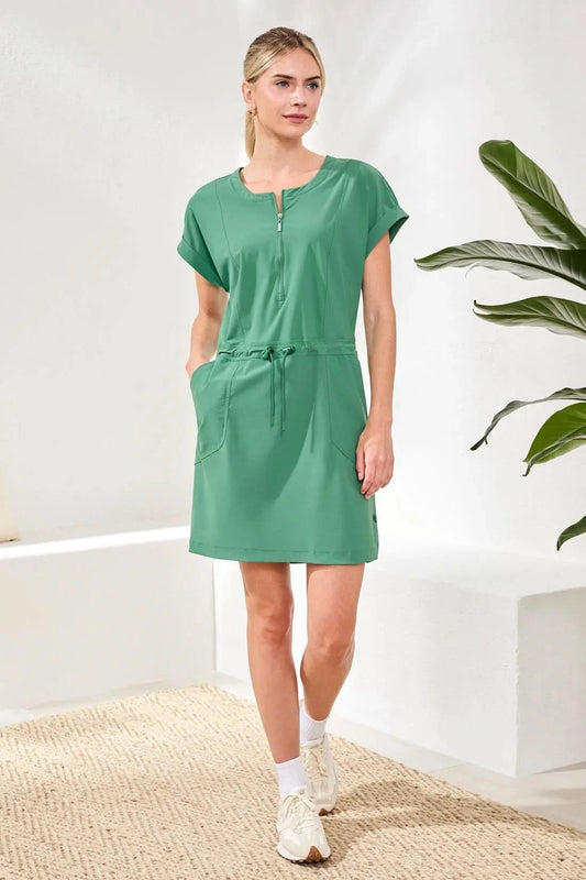A woman stands indoors wearing the Tribal ZIP DRESS, a sporty green zip-front style with a drawcord waist, paired with white socks and sneakers. A green plant is in the background.