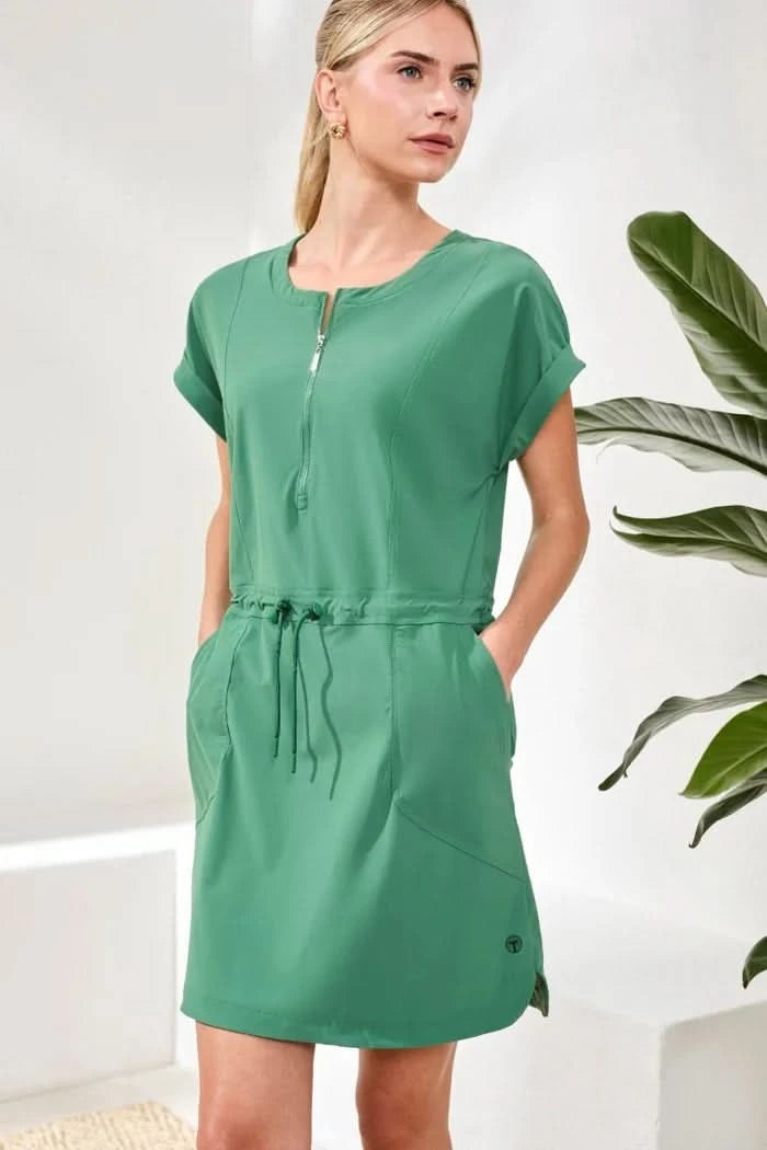 A woman stands indoors wearing the Tribal ZIP DRESS, a sporty green zip-front style with a drawcord waist, paired with white socks and sneakers. A green plant is in the background.