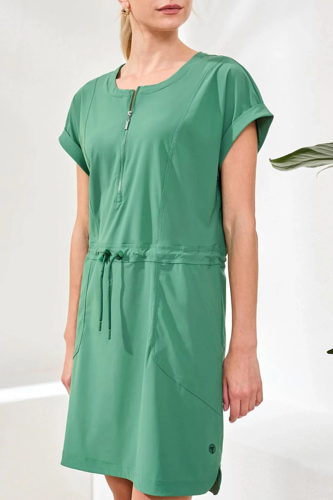 A woman stands indoors wearing the Tribal ZIP DRESS, a sporty green zip-front style with a drawcord waist, paired with white socks and sneakers. A green plant is in the background.