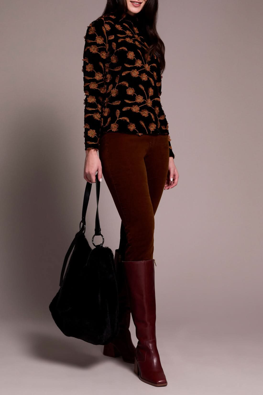 A woman stands against a plain background, dressed in the Tribal MOCK NECK TOP with a black and brown floral pattern, brown pants, Gingerspice burgundy knee-high boots, and carrying a large black bag.