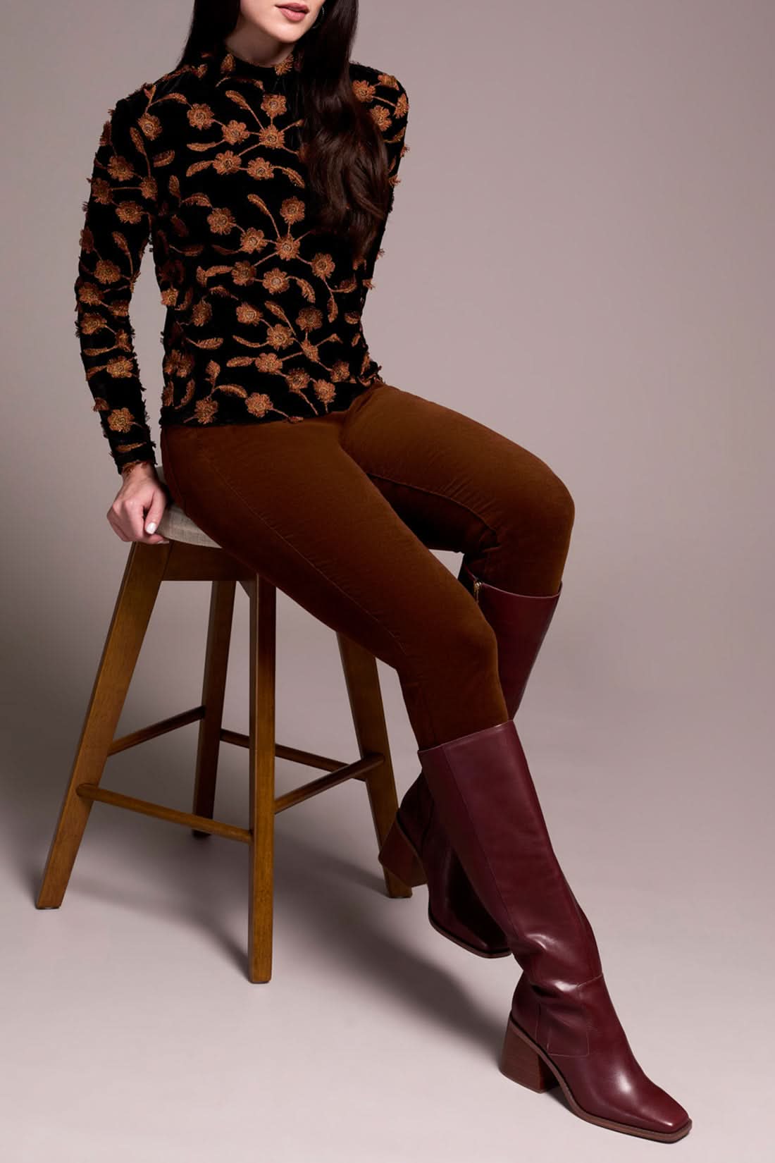 A woman stands against a plain background, dressed in the Tribal MOCK NECK TOP with a black and brown floral pattern, brown pants, Gingerspice burgundy knee-high boots, and carrying a large black bag.
