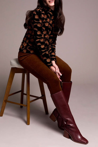 A woman stands against a plain background, dressed in the Tribal MOCK NECK TOP with a black and brown floral pattern, brown pants, Gingerspice burgundy knee-high boots, and carrying a large black bag.