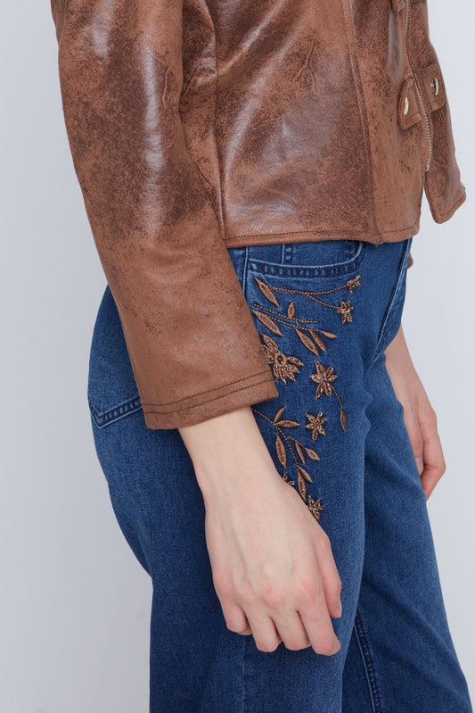 Someone stands against a plain light background wearing CoCo Y Club's Woven Jeans in a mid blue wash with brown floral embroidery, paired with a brown leather jacket.