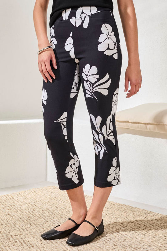 Someone stands indoors on a woven rug wearing Tribal's PULL ON CAPRI W/ POCKETS, styled with a black top, black flats, and a bracelet.