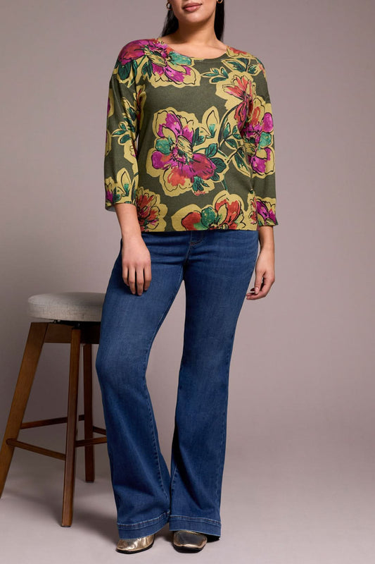 A woman stands beside a wooden stool, wearing the Tribal 3/4 sleeve sweater in Chive and blue flared jeans with silver shoes.