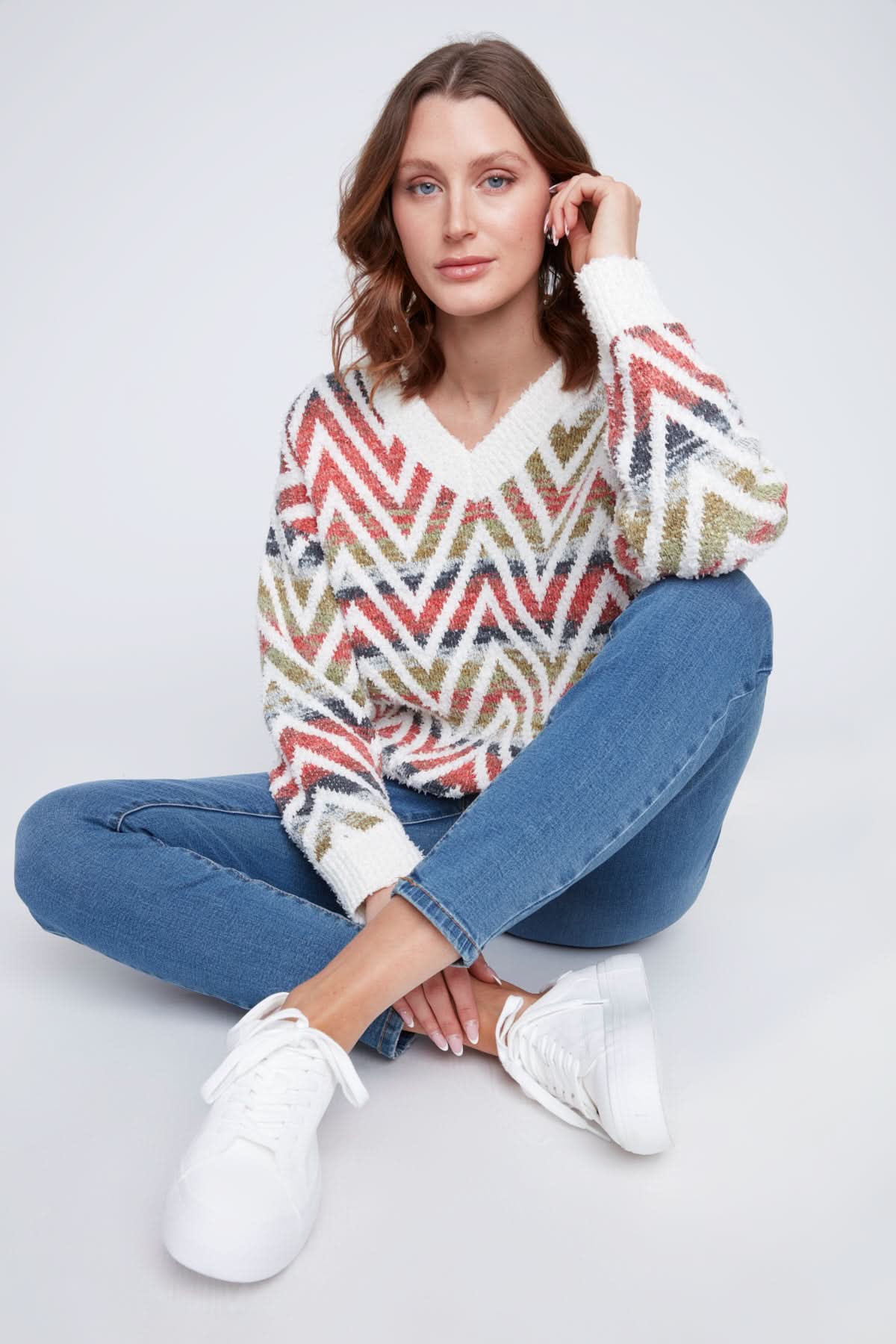 A woman in a CoCo Y Club V neck knitted Sweater with a multicolored zigzag pattern stands with one arm crossed, the other touching her chin, and pairs the cozy piece with blue jeans.