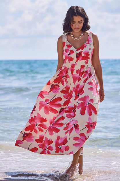 A woman wearing the Tribal Sleeveless Flow Maxi Dress, featuring large red floral prints and a stretch back panel, stands at the water’s edge, gazing down as gentle waves touch her feet.
