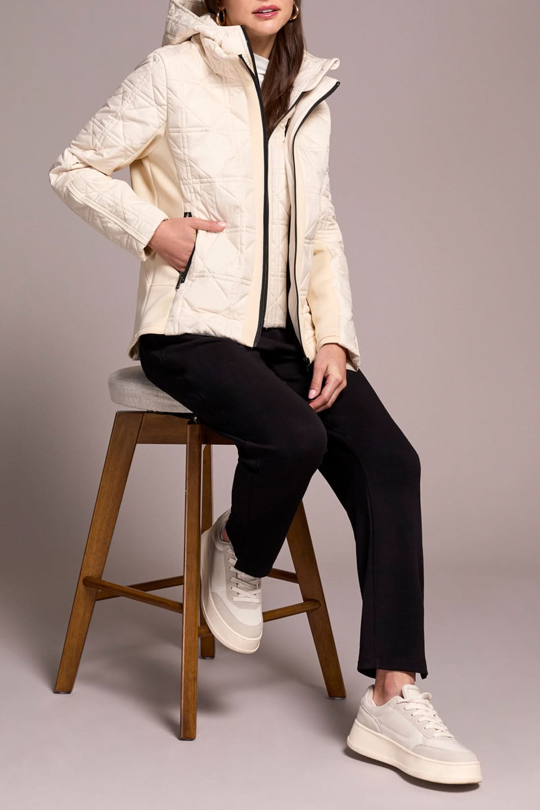 A person wears the Tribal Puffer W/Removable Hood in cream, paired with a white turtleneck, black pants, and white sneakers, standing against a plain background.