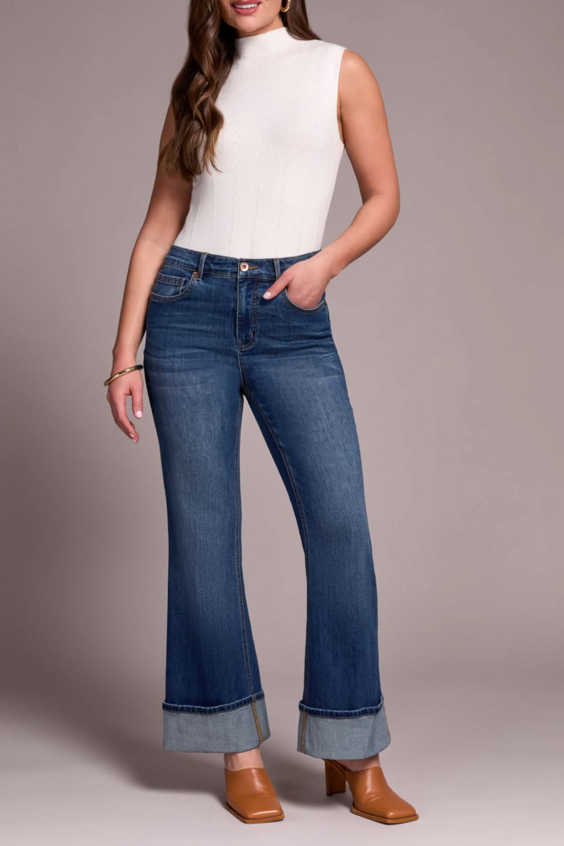 A woman stands with one hand in her pocket, wearing a sleeveless white turtleneck, Tribal BROOKE PALAZZO JEANS in antique blue, and tan heeled shoes.
