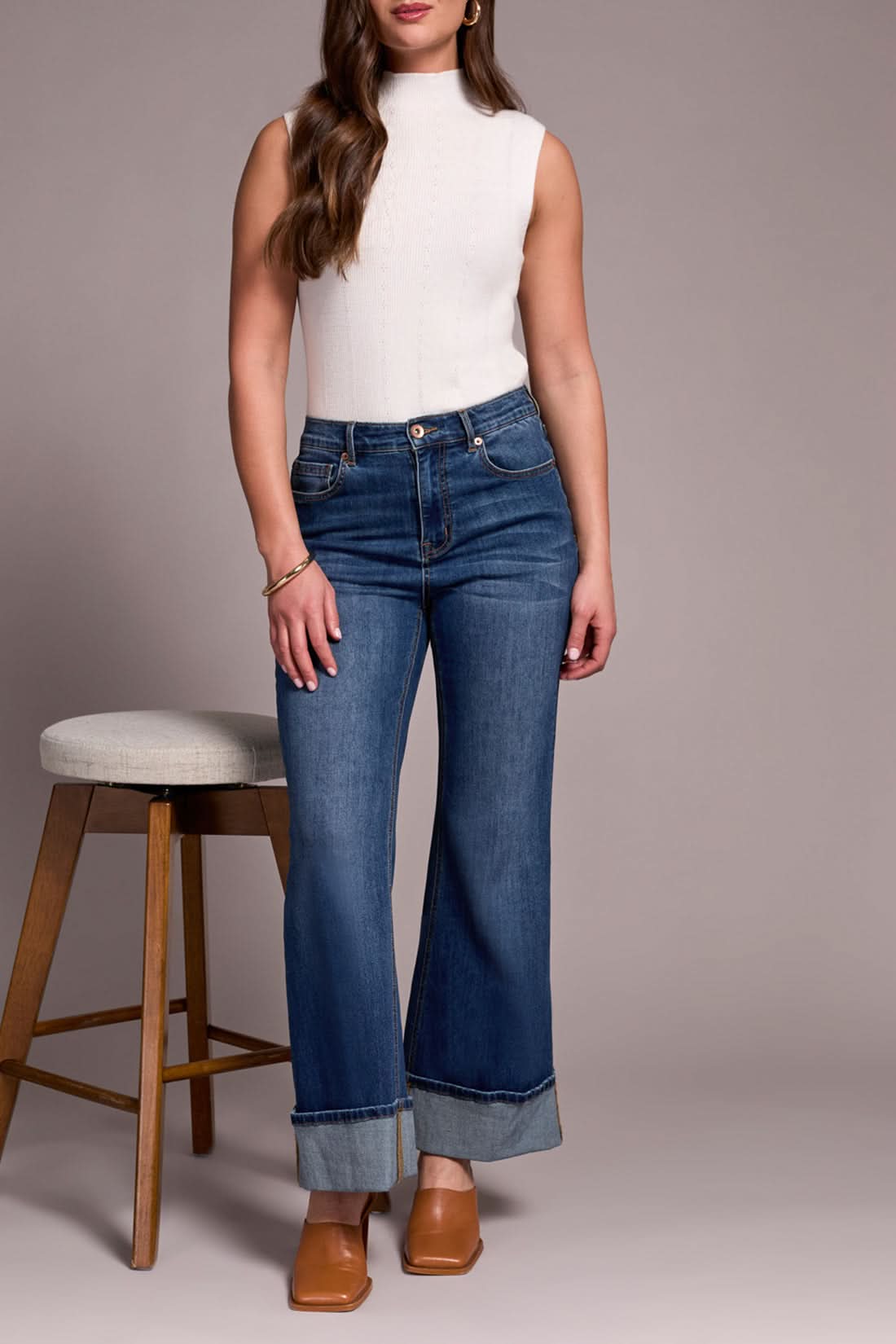 A woman stands with one hand in her pocket, wearing a sleeveless white turtleneck, Tribal BROOKE PALAZZO JEANS in antique blue, and tan heeled shoes.