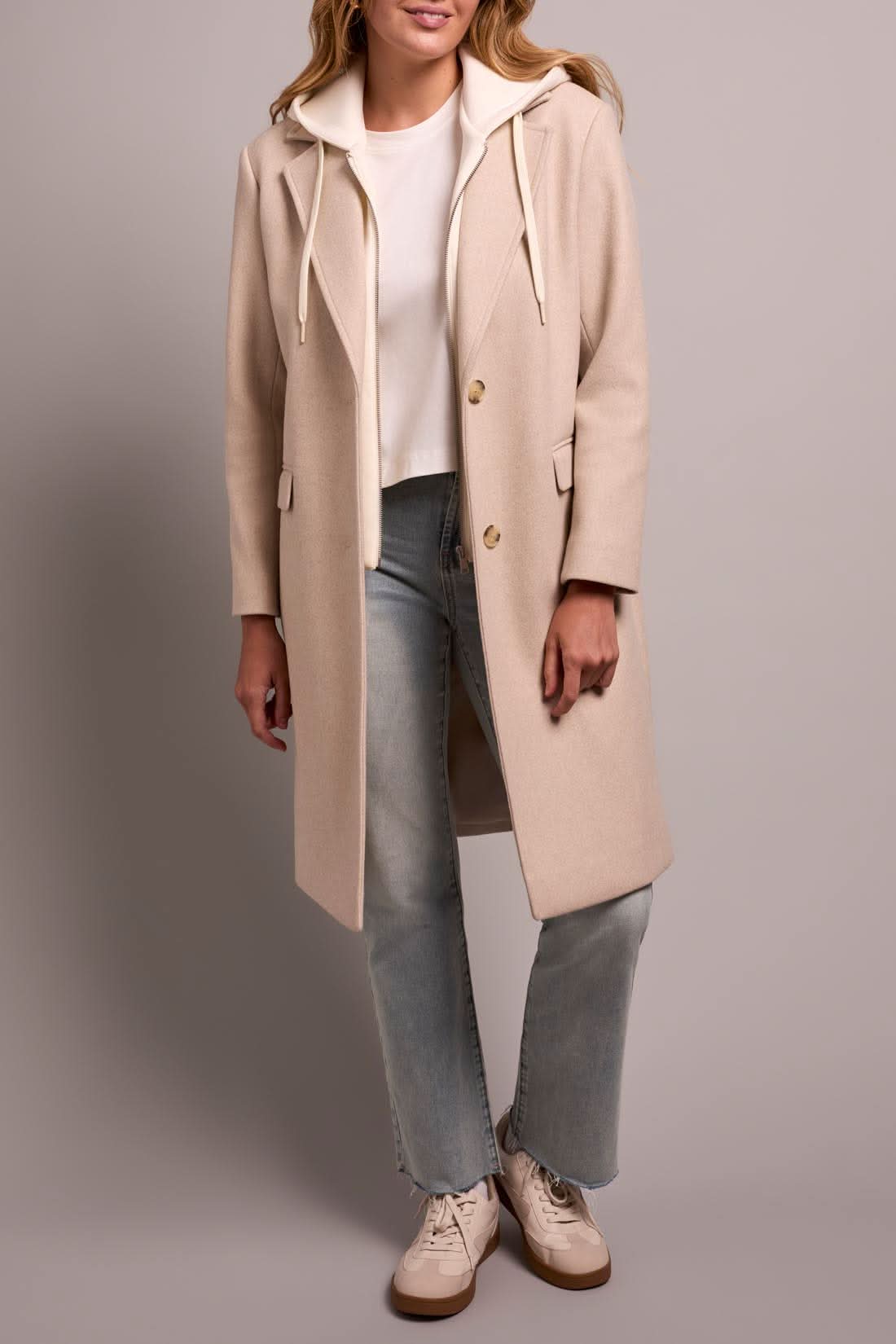 A person models the Tribal TRENCH COAT W/HOODIE, paired with light blue jeans and white sneakers, against a plain background.