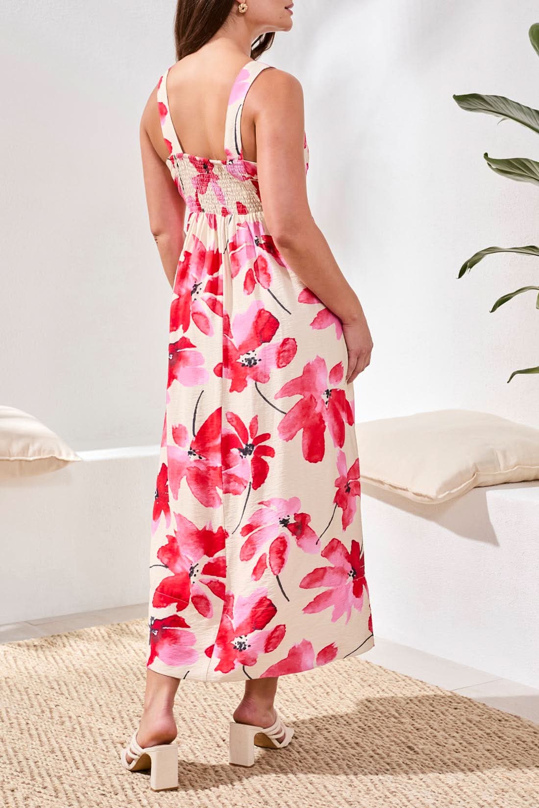 A woman wearing the Tribal Sleeveless Flow Maxi Dress, featuring large red floral prints and a stretch back panel, stands at the water’s edge, gazing down as gentle waves touch her feet.