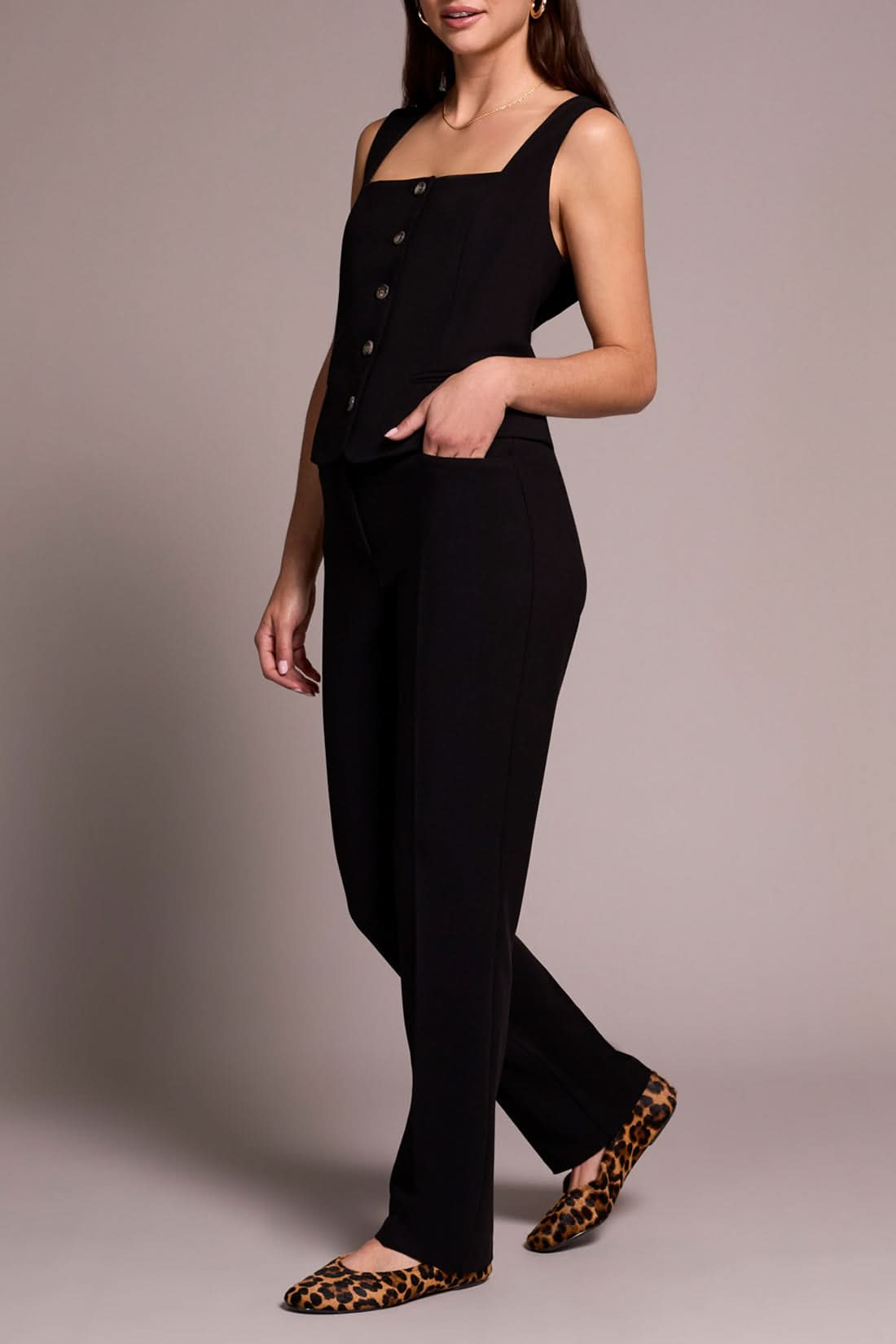 A person stands smiling in a sleeveless black buttoned top, Tribal FLY FRT TROUSER black pants, and animal print flats against a neutral background.