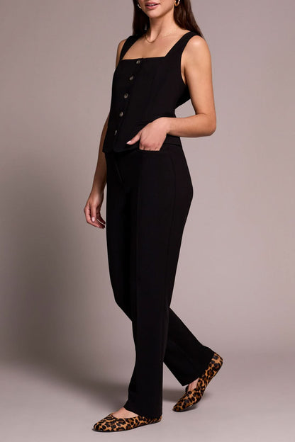 A person stands smiling in a sleeveless black buttoned top, Tribal FLY FRT TROUSER black pants, and animal print flats against a neutral background.