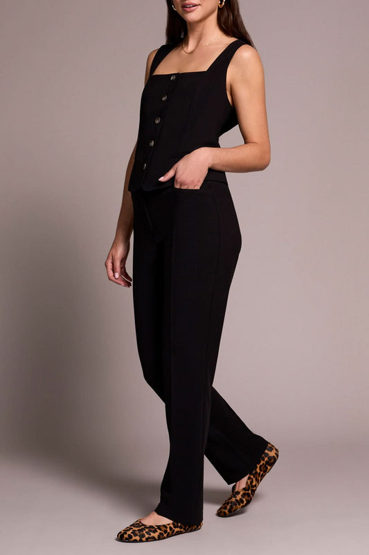 A person stands smiling in a sleeveless black buttoned top, Tribal FLY FRT TROUSER black pants, and animal print flats against a neutral background.