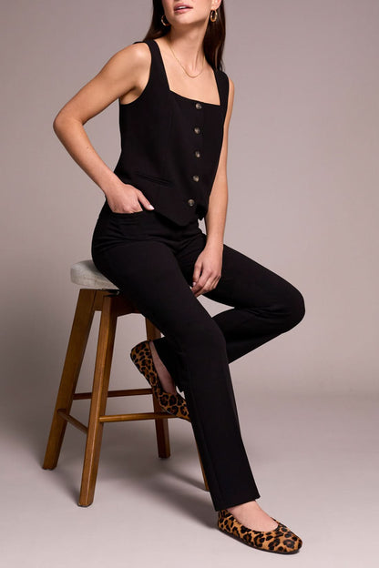 A person stands smiling in a sleeveless black buttoned top, Tribal FLY FRT TROUSER black pants, and animal print flats against a neutral background.