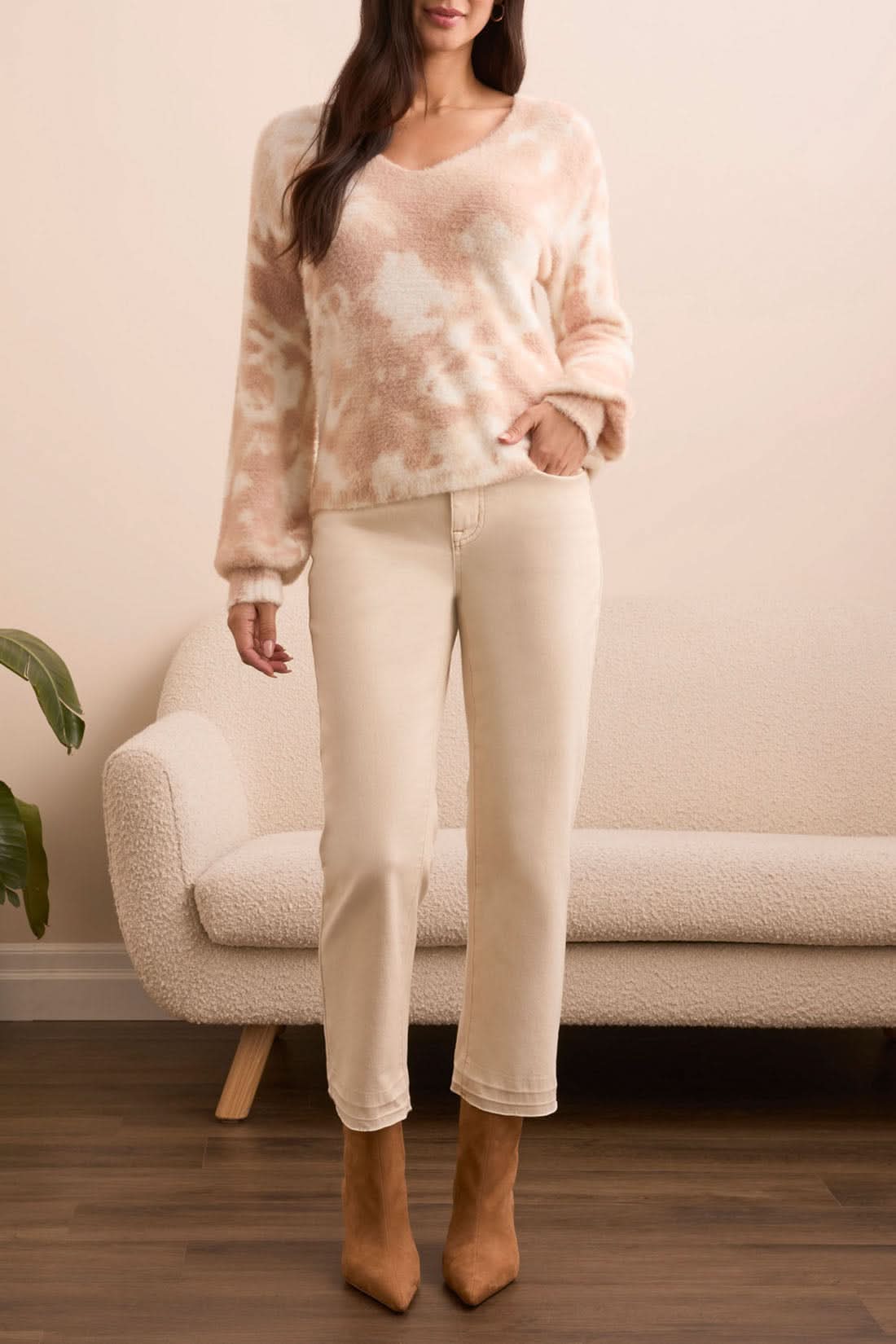 A woman stands indoors in front of a light sofa and plant, wearing Tribal’s WEAR 2 WAYS SWEATER in pink and white, paired with light beige jeans and tan boots.