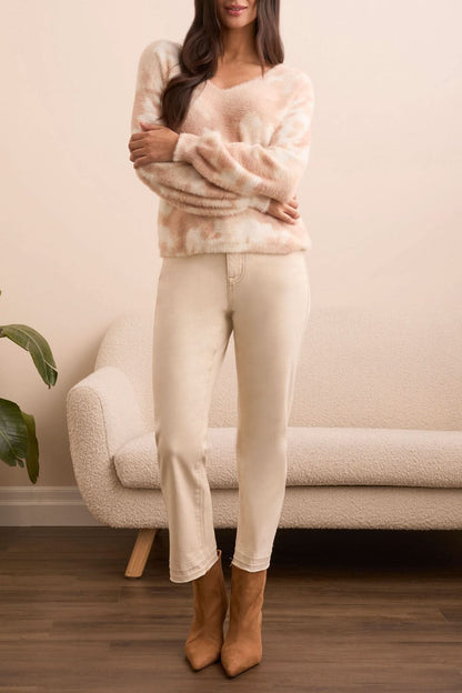 A woman stands indoors in front of a light sofa and plant, wearing Tribal’s WEAR 2 WAYS SWEATER in pink and white, paired with light beige jeans and tan boots.