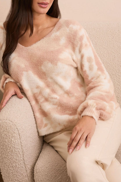 A woman stands indoors in front of a light sofa and plant, wearing Tribal’s WEAR 2 WAYS SWEATER in pink and white, paired with light beige jeans and tan boots.