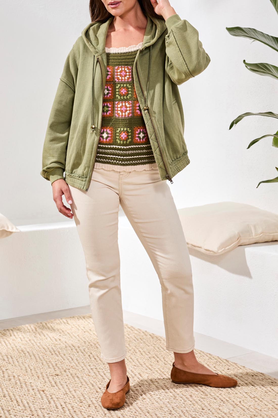 A woman stands indoors wearing the Tribal Puff Sleeve Hoodie in light green, zipped over a colorful crocheted top and paired with beige pants.