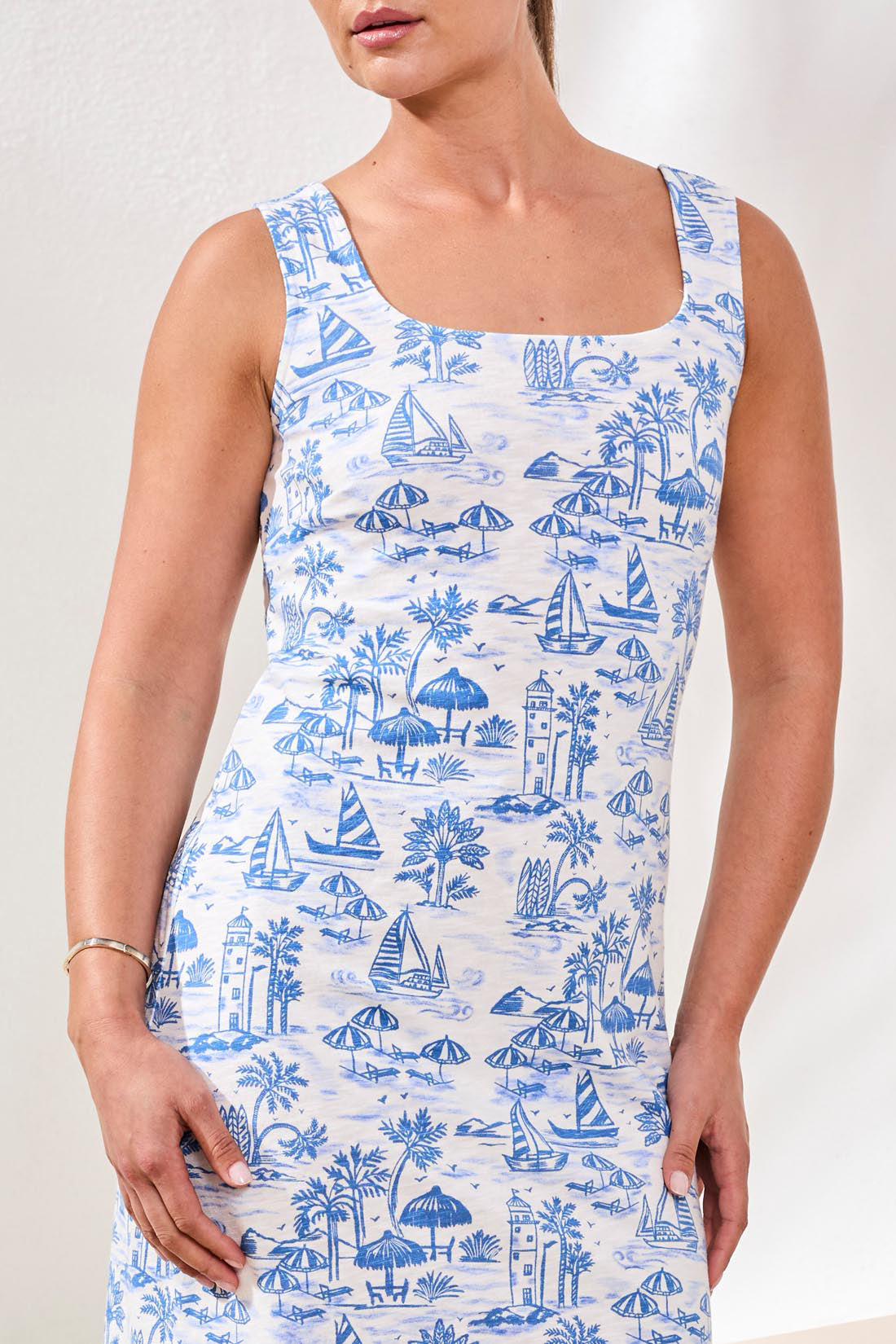 A woman in a Tribal Sleeveless Square Neck Dress stands indoors on a woven rug, wearing beige wedge sandals and holding a white handbag.