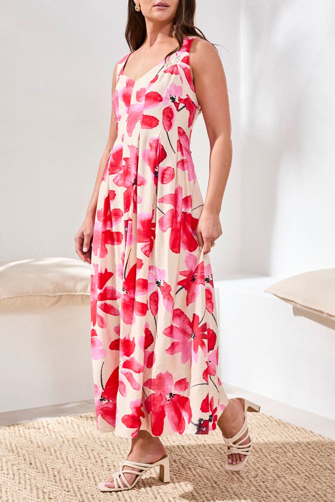 A woman wearing the Tribal Sleeveless Flow Maxi Dress, featuring large red floral prints and a stretch back panel, stands at the water’s edge, gazing down as gentle waves touch her feet.