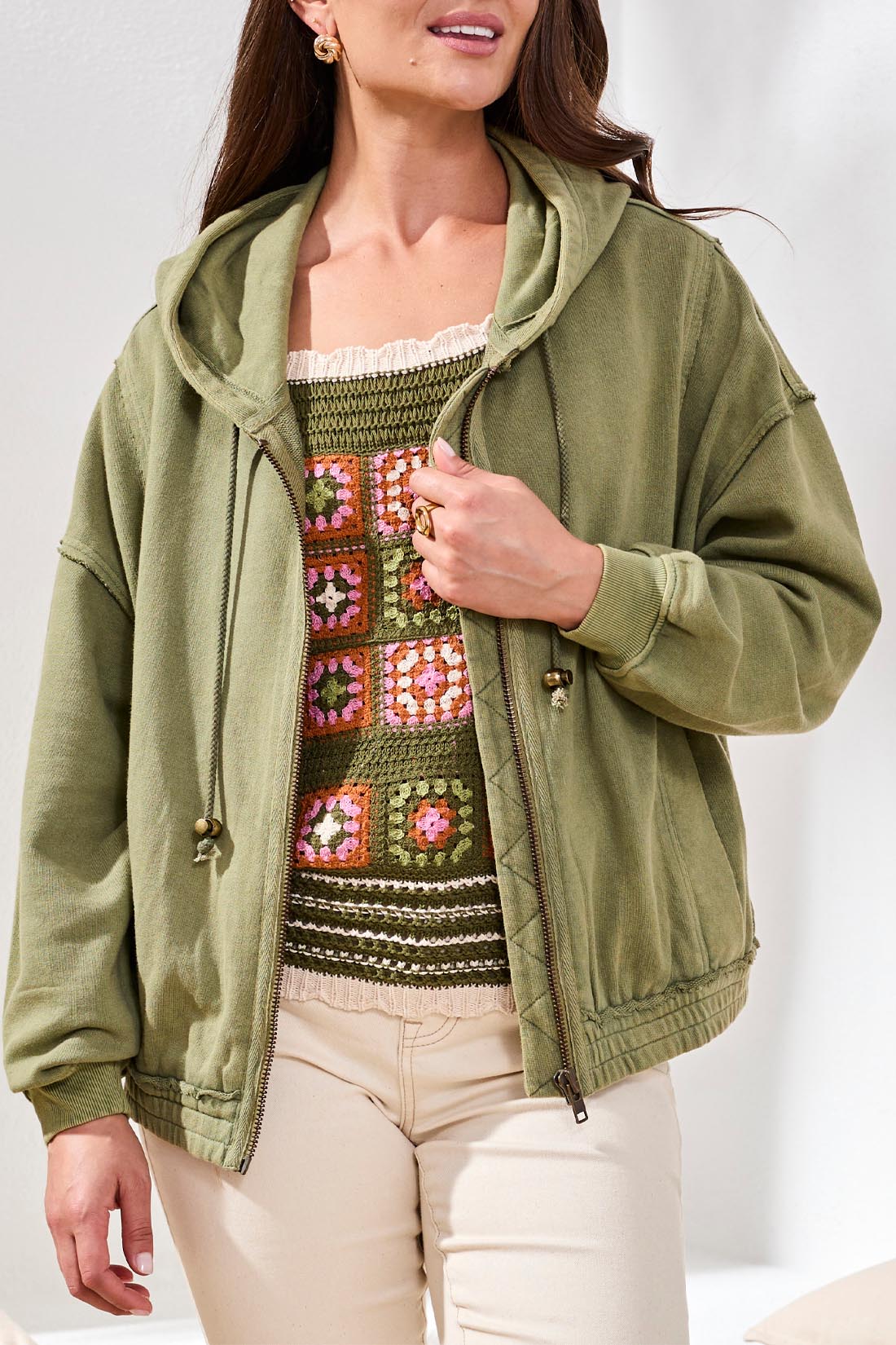 A woman stands indoors wearing the Tribal Puff Sleeve Hoodie in light green, zipped over a colorful crocheted top and paired with beige pants.