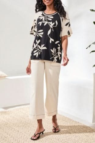A woman stands indoors wearing Tribal's FLUTTER SLEEVE CREW NECK, a black and beige leaf-patterned top made from a rayon spandex blend, paired with beige wide-leg pants and black sandals.