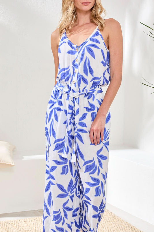 A woman models the Tribal Blue Flowy Jumpsuit, a sleeveless cotton style with blue leaf prints, button front, and a tied waist, standing indoors against a light background to showcase its chic silhouette.
