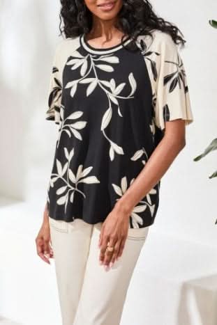 A woman stands indoors wearing Tribal's FLUTTER SLEEVE CREW NECK, a black and beige leaf-patterned top made from a rayon spandex blend, paired with beige wide-leg pants and black sandals.