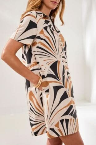 A woman poses indoors in the Tribal Placket Dress, featuring short sleeves and a beige, black, and brown leaf print. Styled as a summer look with black slide sandals, she smiles with one hand at her side.