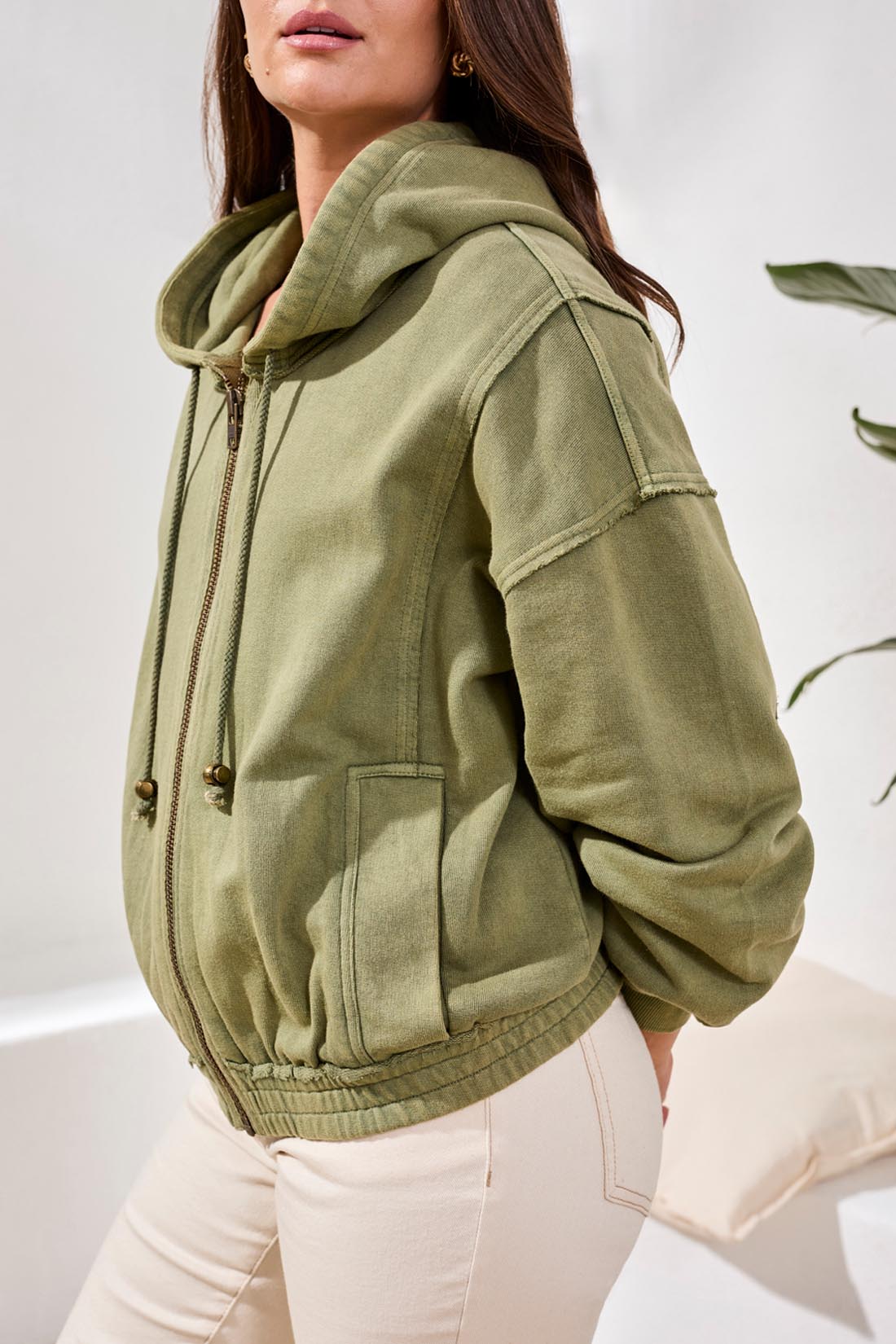 A woman stands indoors wearing the Tribal Puff Sleeve Hoodie in light green, zipped over a colorful crocheted top and paired with beige pants.