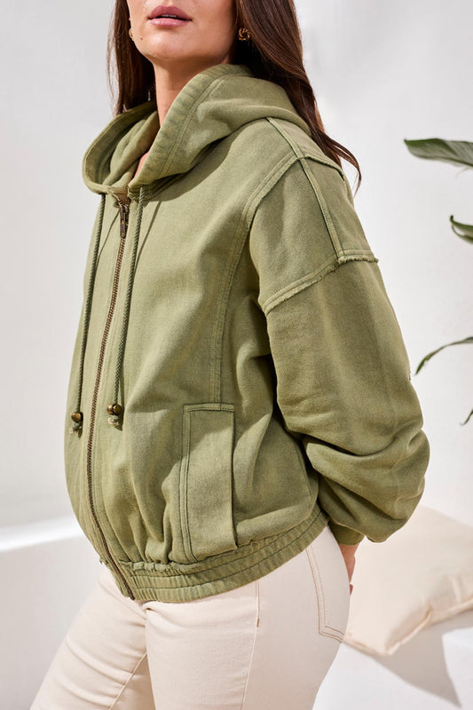 A woman stands indoors wearing the Tribal Puff Sleeve Hoodie in light green, zipped over a colorful crocheted top and paired with beige pants.