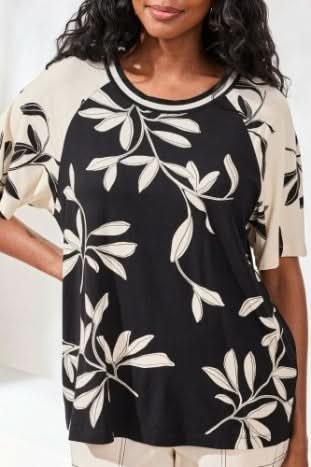 A woman stands indoors wearing Tribal's FLUTTER SLEEVE CREW NECK, a black and beige leaf-patterned top made from a rayon spandex blend, paired with beige wide-leg pants and black sandals.