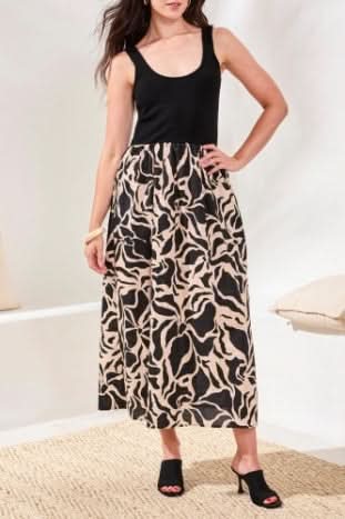 A woman wears the Tribal SLEEVELESS TWOFER DRESS, featuring a black top and an A-line beige and black abstract cotton skirt, styled with black open-toe heeled shoes.