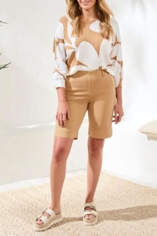 A woman stands indoors on a woven rug, wearing a white and tan abstract patterned top, Tribal Comfort Stretch Pull on Shorts in tan, and white sandals.