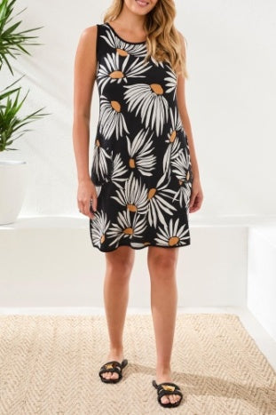 A woman models the Tribal REVERSIBLE A-LINE DRESS—sleeveless, black, with a white and yellow daisy print—standing indoors by a plant. This versatile dress is perfect for travel or everyday wear.