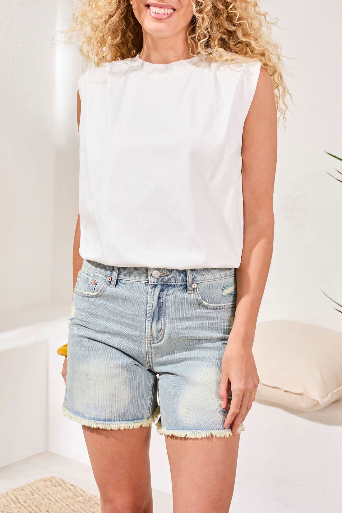 A woman in the Tribal Bubble Tank Top, light denim shorts, white sandals, and a yellow bangle stands indoors on a woven rug beside white cushions and a potted plant.