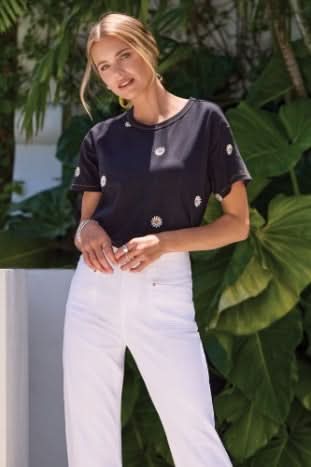 A woman wearing the Tribal EMBROIDERED CREW NECK TOP with black fabric and small white daisy prints pairs it with a white skirt as she sits on a white surface, smiling.