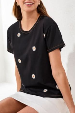 A woman wearing the Tribal EMBROIDERED CREW NECK TOP with black fabric and small white daisy prints pairs it with a white skirt as she sits on a white surface, smiling.