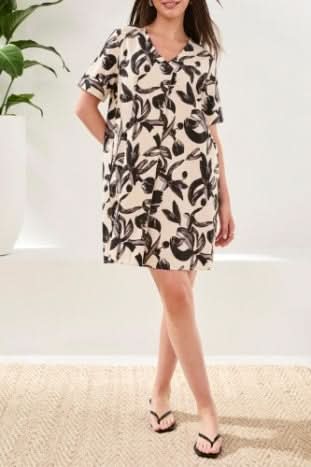 A woman stands indoors wearing the Tribal TUNIC DRESS, a breathable cotton, knee-length cream summer dress with black leafy print and short sleeves, paired with black flip-flops.