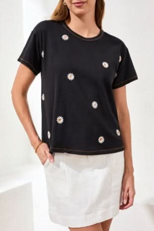 A woman wearing the Tribal EMBROIDERED CREW NECK TOP with black fabric and small white daisy prints pairs it with a white skirt as she sits on a white surface, smiling.